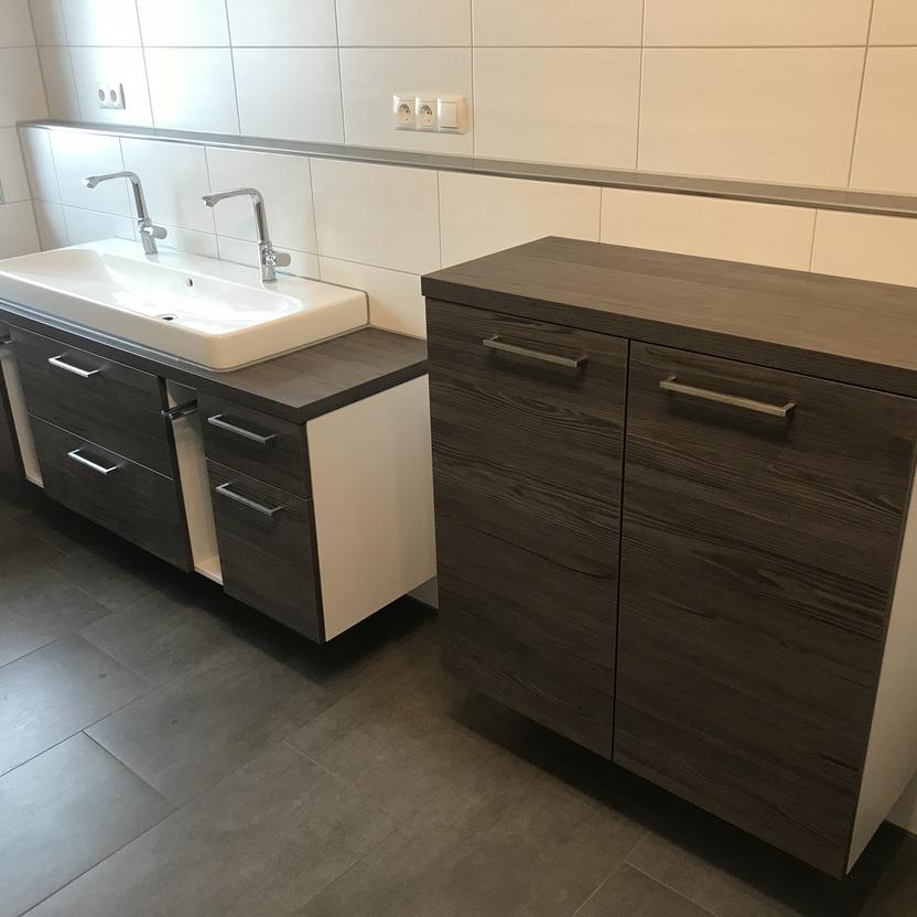 Doppelwaschbecken mit braunen Unterschränken und Spiegel an einer gefliesten Wand.