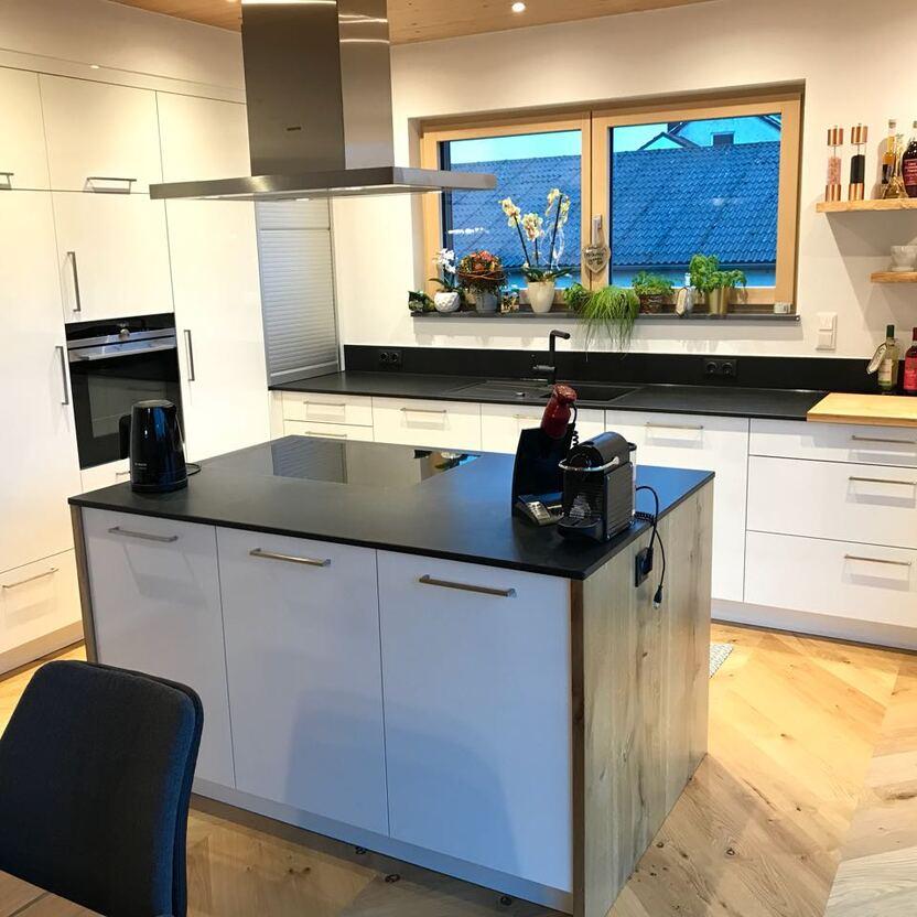 Küchenmöbel mit Kochinsel, weißen Fronten, schwarzer Arbeitsplatte und offenen Holzregalen unter dem Fenster