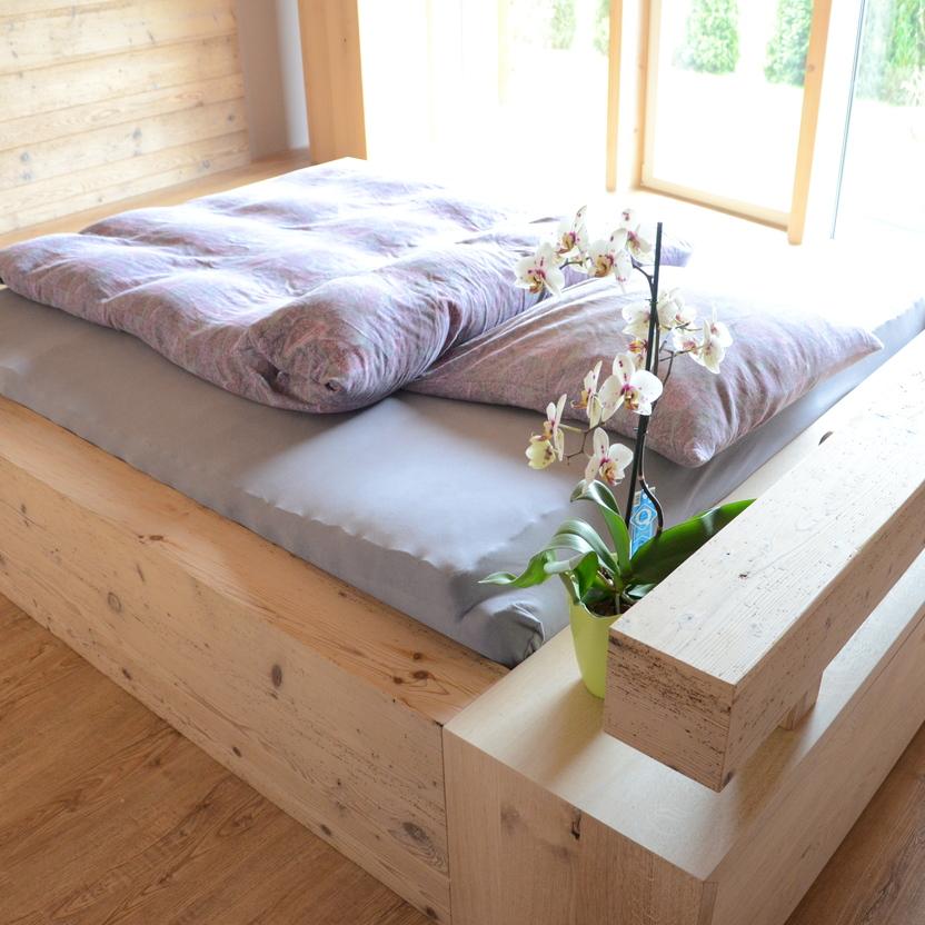 Ein minimalistisches Holzbett mit grauer Bettwäsche und einer Orchidee in einem sonnendurchfluteten Raum.