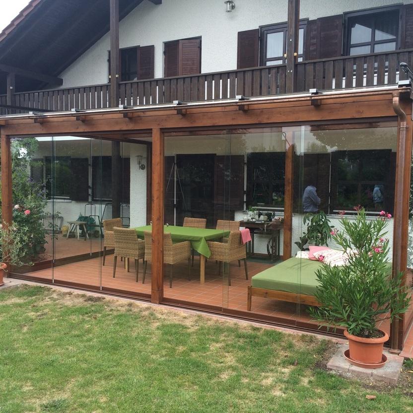 Holzpergola mit Glasfront und gemütlicher Sitzecke, angrenzend an ein Haus mit Garten.