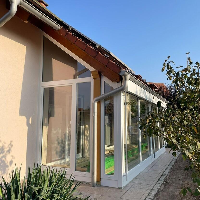 Wintergarten mit weißem Kunststoffrahmen und bodentiefen Fenstern entlang der Hausfassade bei sonnigem Wetter