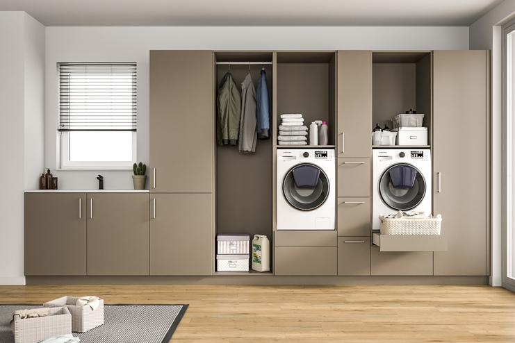 Ein heller Raum mit einem großen, maßgeschneiderten Schranksystem in Taupe, integrierten Waschmaschinen, Ablageflächen und einem Fenster mit Jalousien.
